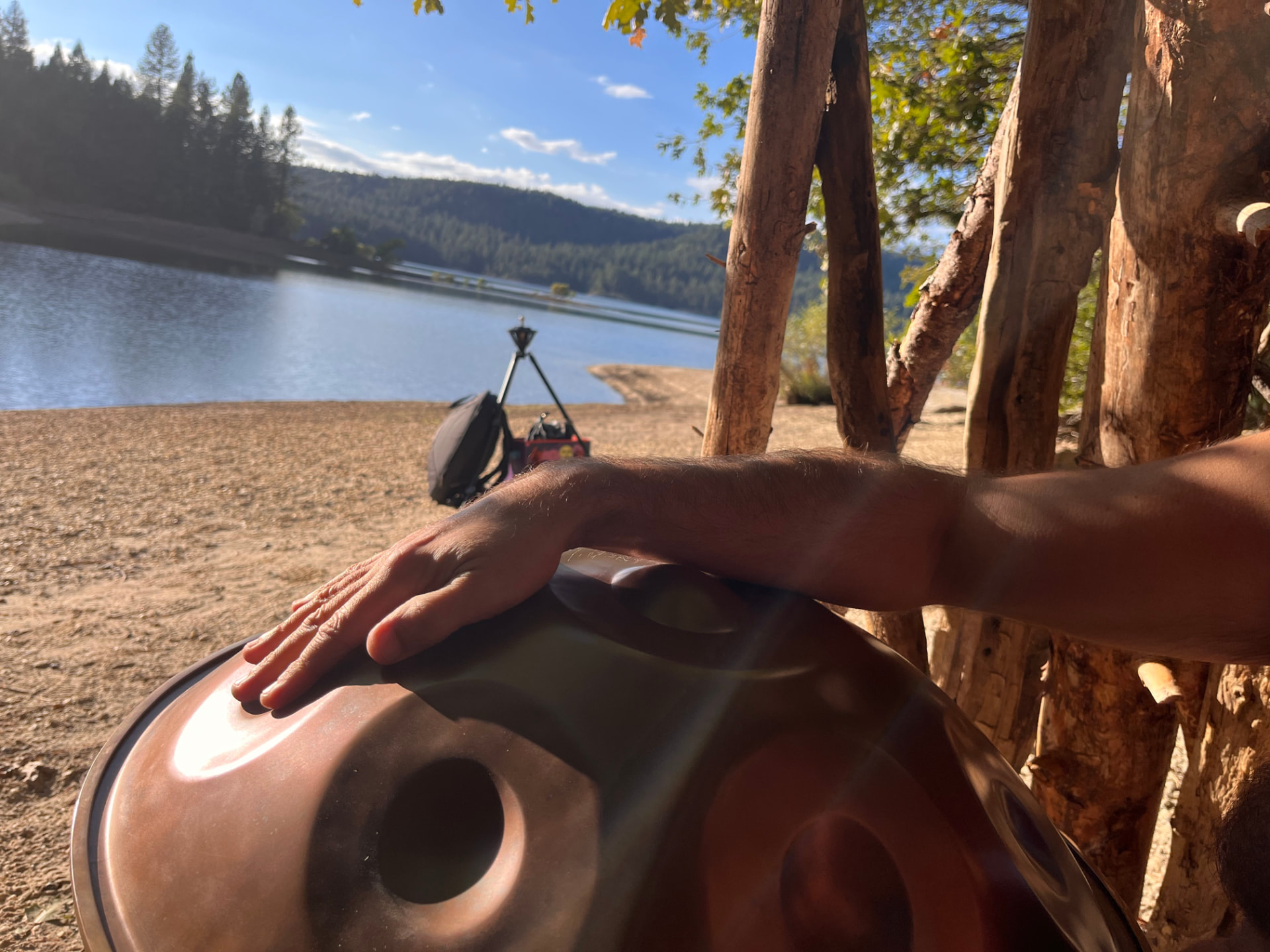 A hand playing a handpan beside a sunlit mountain lake